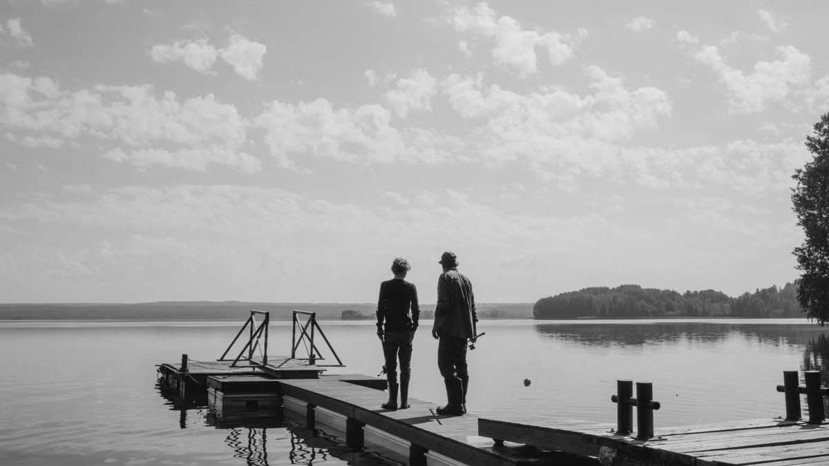 Two people stand on a lakeside pier under a cloudy sky, reflecting a peaceful atmosphere.